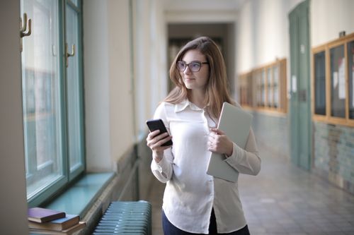 Free Female student with glasses holding a phone and laptop near window in school hallway. Stock Photo
