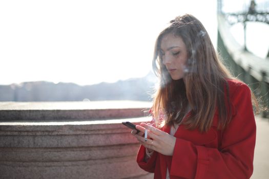 阳光明媚的日子里，一位穿着红色外套的女子在外面抽烟、发短信