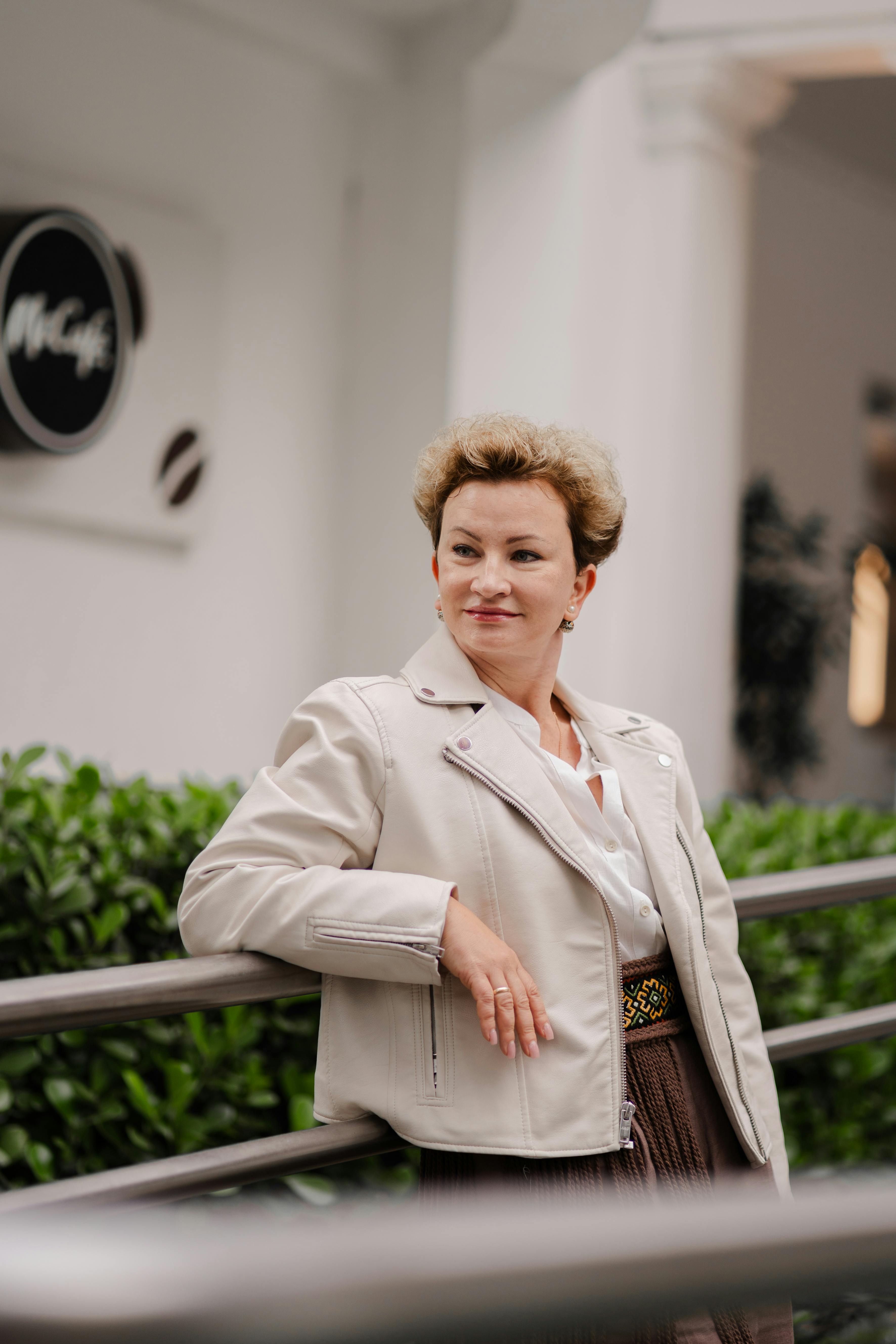 A confident woman posing outdoors, wearing a beige jacket and stylish attire, with a McCafe sign in the background.
