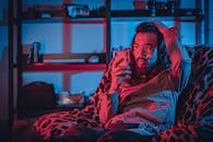 Surprised adult unshaven ethnic male in eyeglasses with pouting lips browsing internet on cellphone while touching head and sitting on comfortable armchair near shelf in apartment with neon light