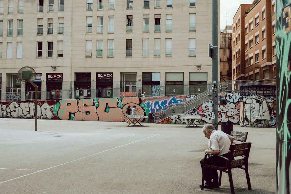Hombre mayor sentado en un banco junto a una cancha de baloncesto cubierta de graffiti en Barcelona.