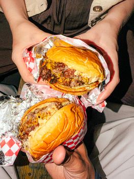 Close-up of two burgers wrapped in foil held by friends, perfect for summer picnics.