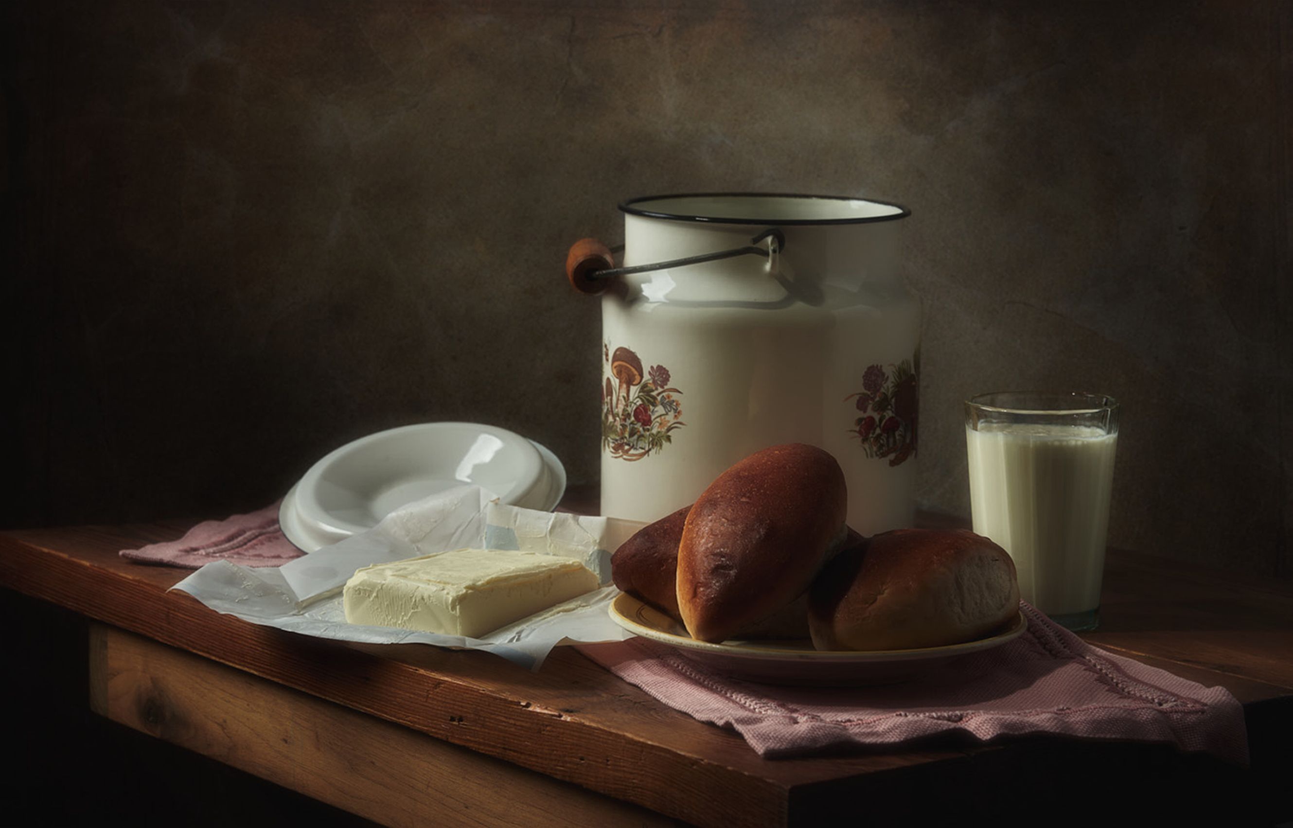 Cozy still life featuring milk, bread rolls, butter, and an enamel container on a wooden table.