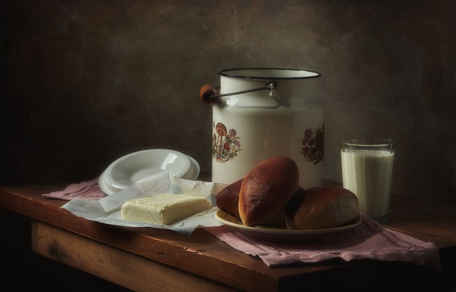 Cozy still life featuring milk, bread rolls, butter, and an enamel container on a wooden table.