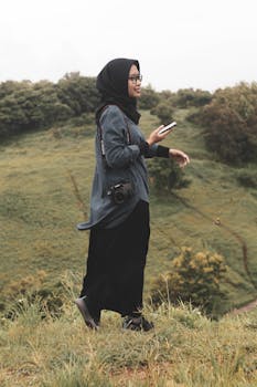 一名戴头巾的妇女手持相机和智能手机，站在绿色的乡村景观中