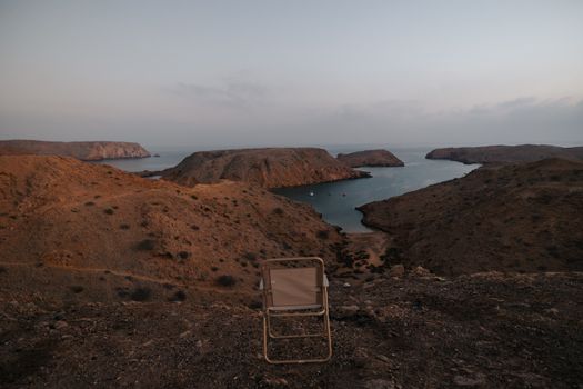 A stunning view of rocky cliffs and a serene bay in Muscat, Oman during twilight with an empty chair in the foreground.