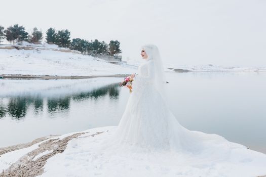A bride in a white gown stands elegantly by a snow-covered lake, holding a bouquet, surrounded by winter scenery.