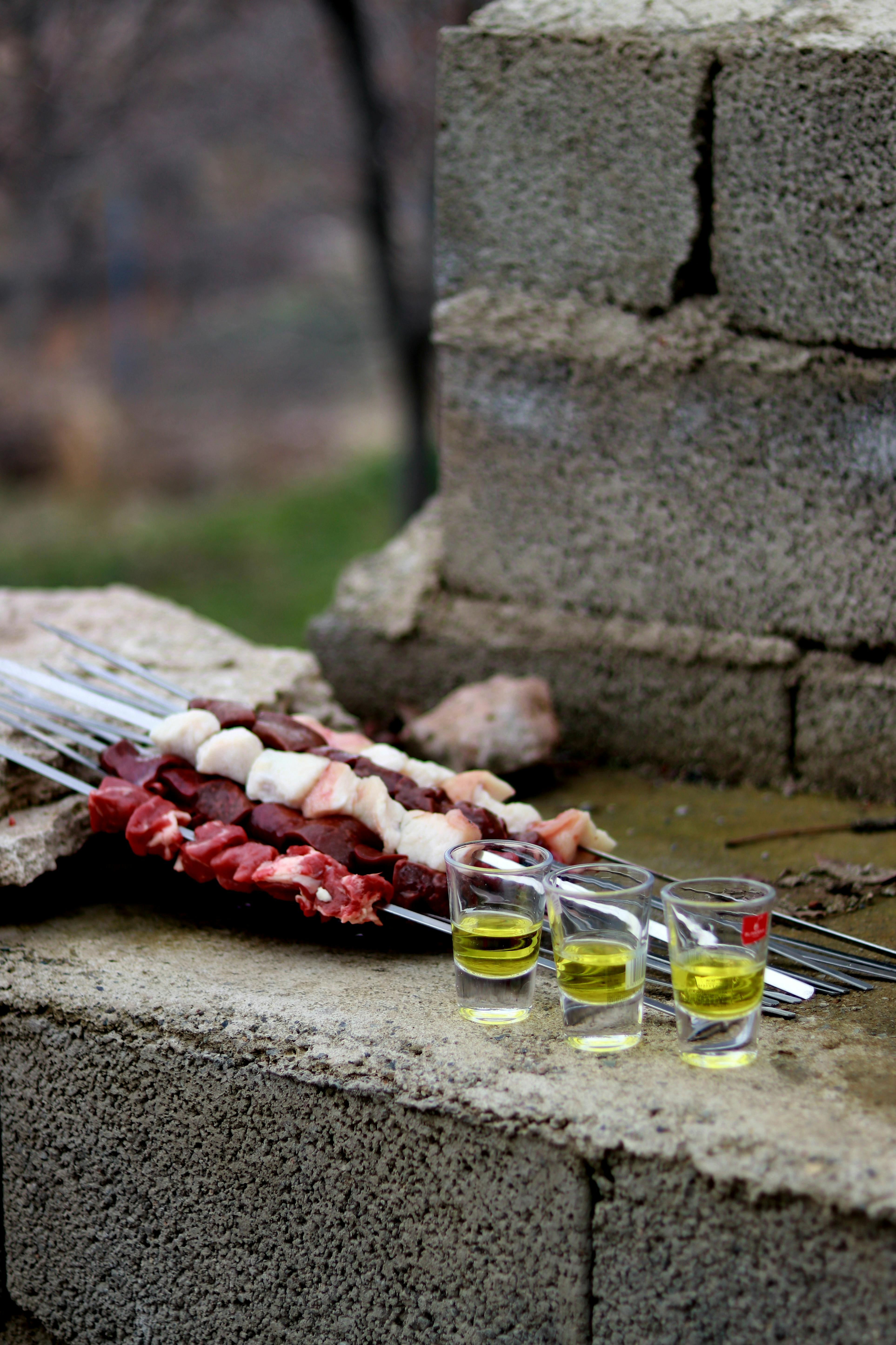 Raw meat skewers on a stone grill with shot glasses, in an outdoor setting.