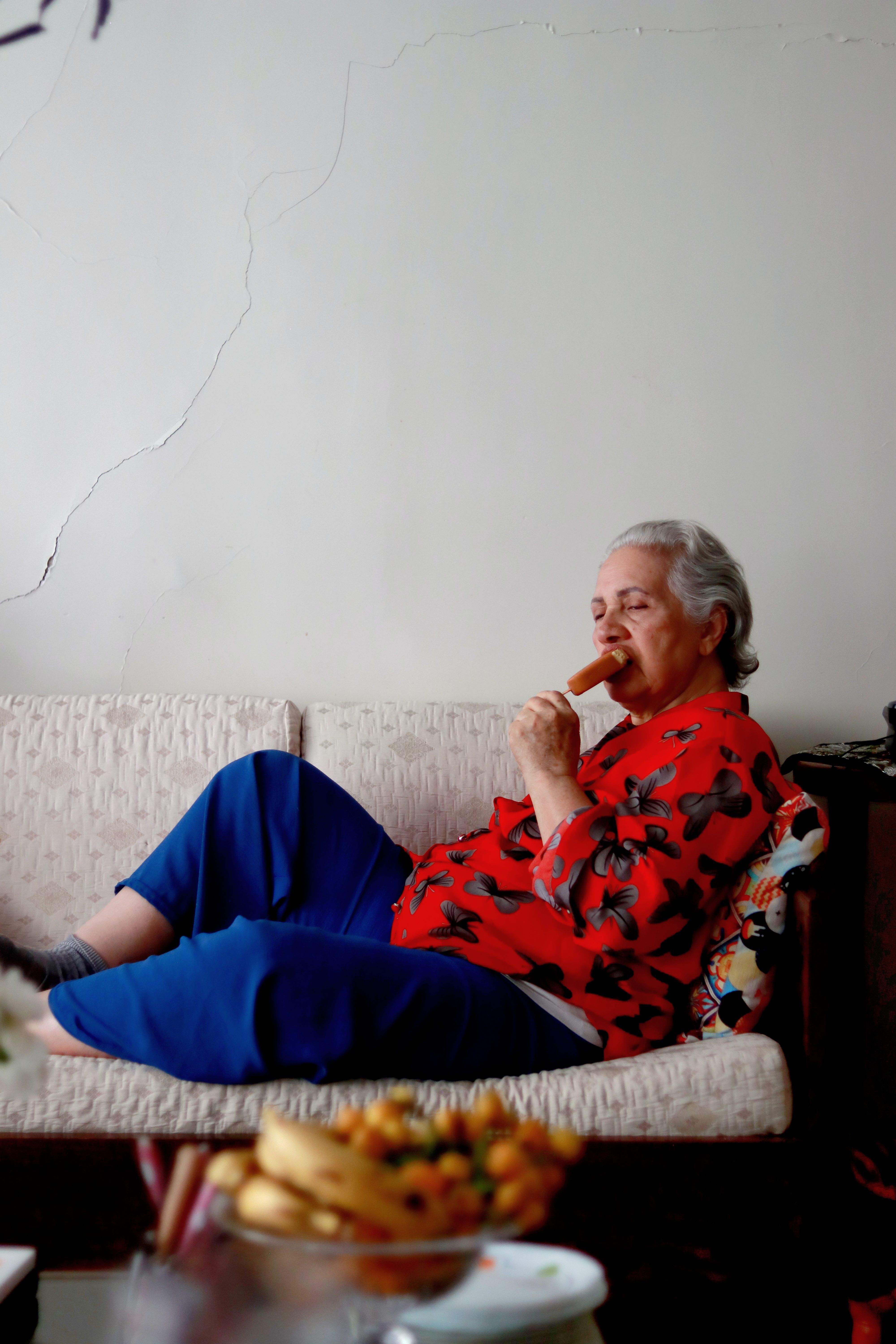 Senior woman in colorful attire relaxing on a couch, savoring a snack indoors.