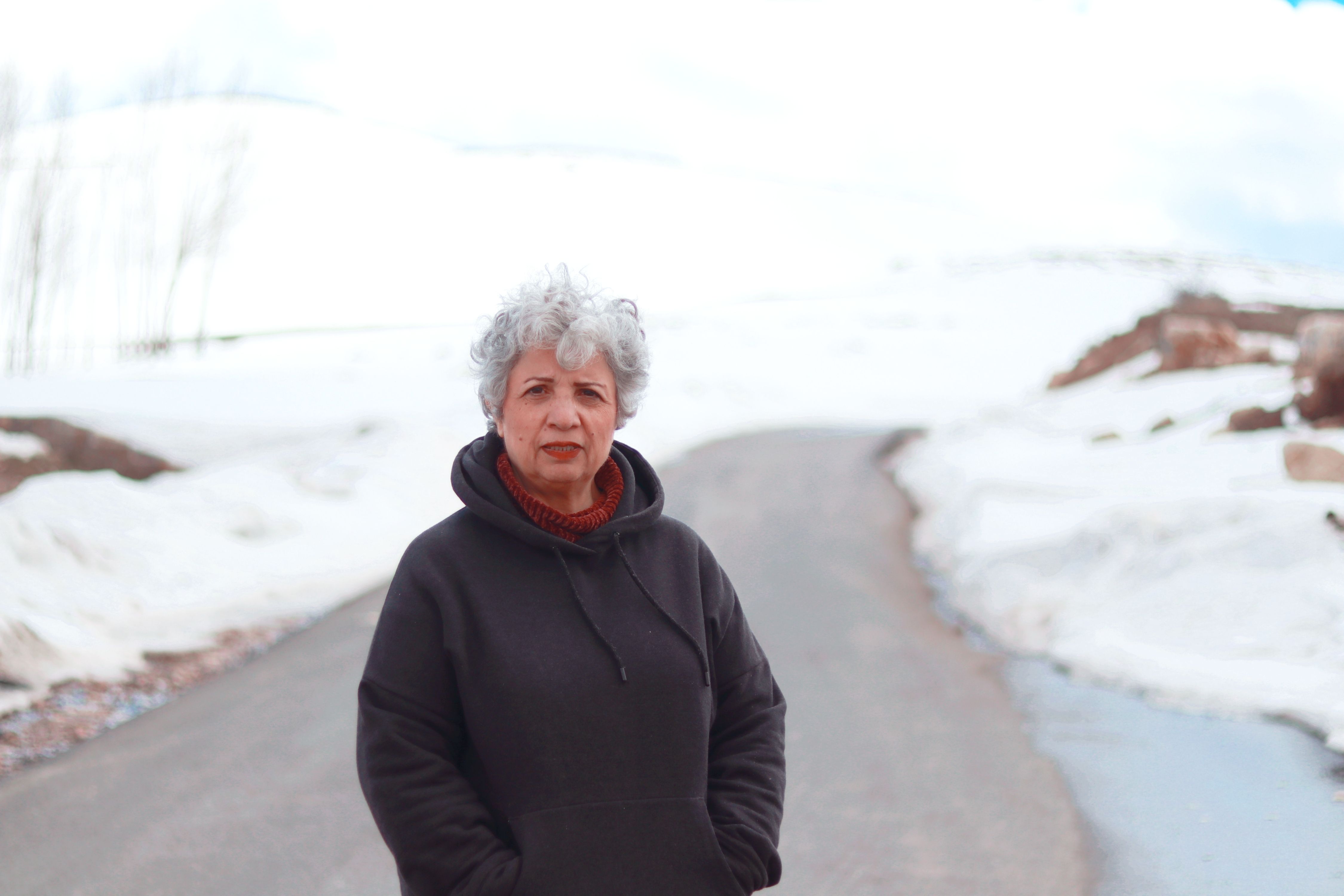 Senior woman standing on a snowy road wearing a dark hoodie, winter scenery.