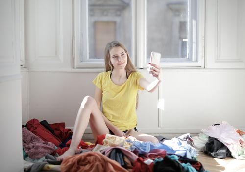 Free Young woman in a yellow top smiling and taking a selfie surrounded by clothes indoors. Stock Photo