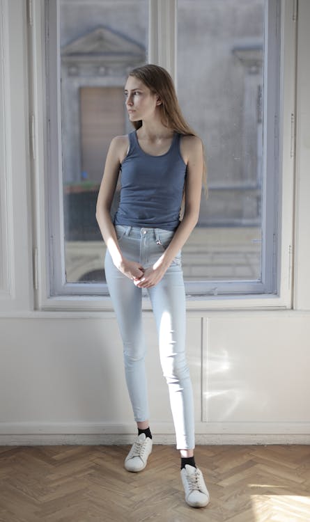 Free Woman in Blue Tank Top and White Pants Standing Beside Window Stock Photo