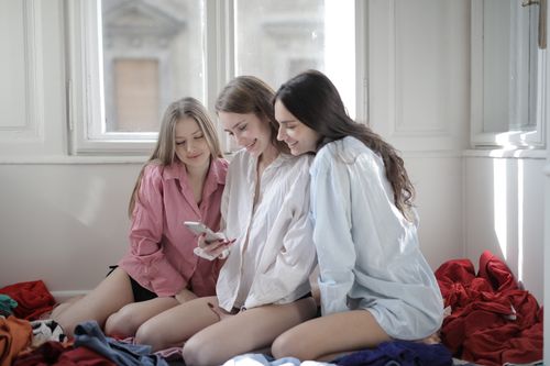 Free Company of friendly females wearing casual shirts sitting on floor in untidy room and messaging on social media while chilling during weekend at home Stock Photo