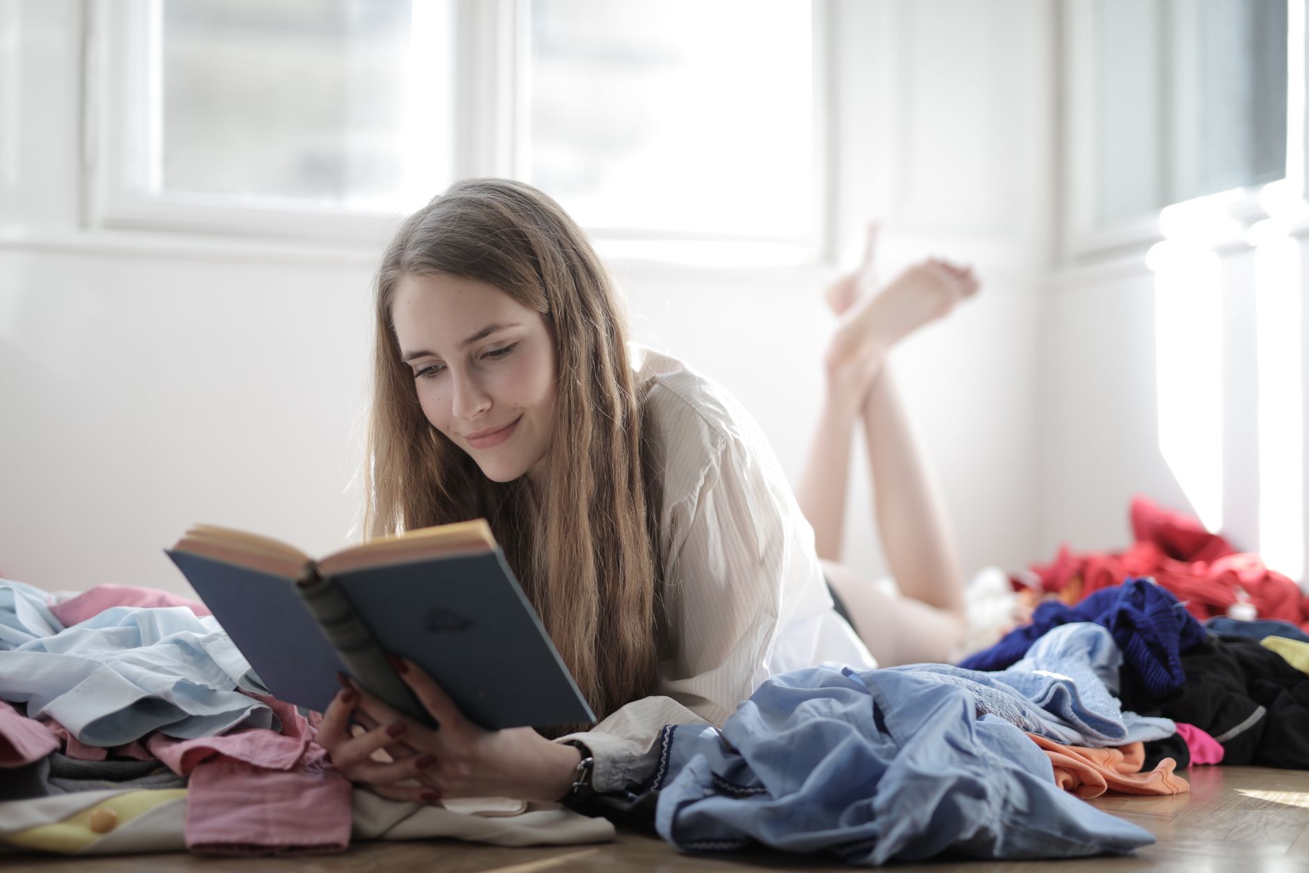 A young woman with long hair enjoys reading a book while lounging on the floor surrounded by clothes.