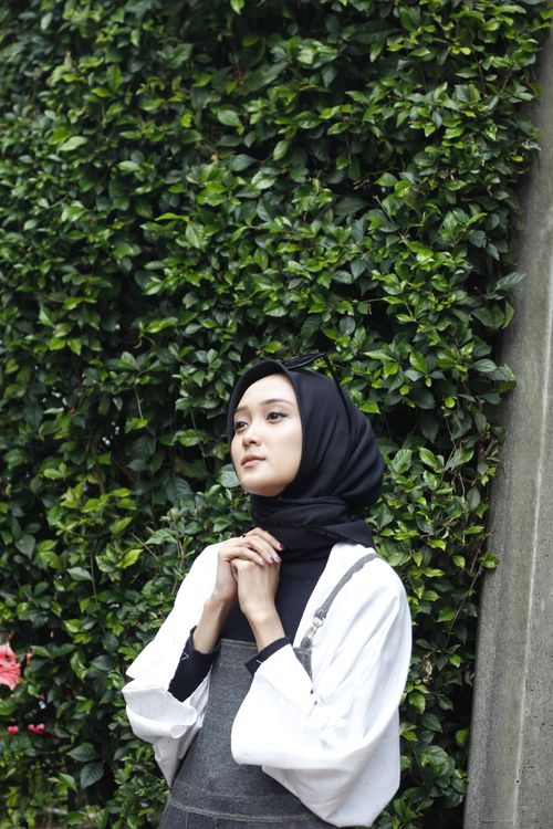 Free Portrait of a young woman wearing a hijab, standing against vibrant green leaves. Stock Photo