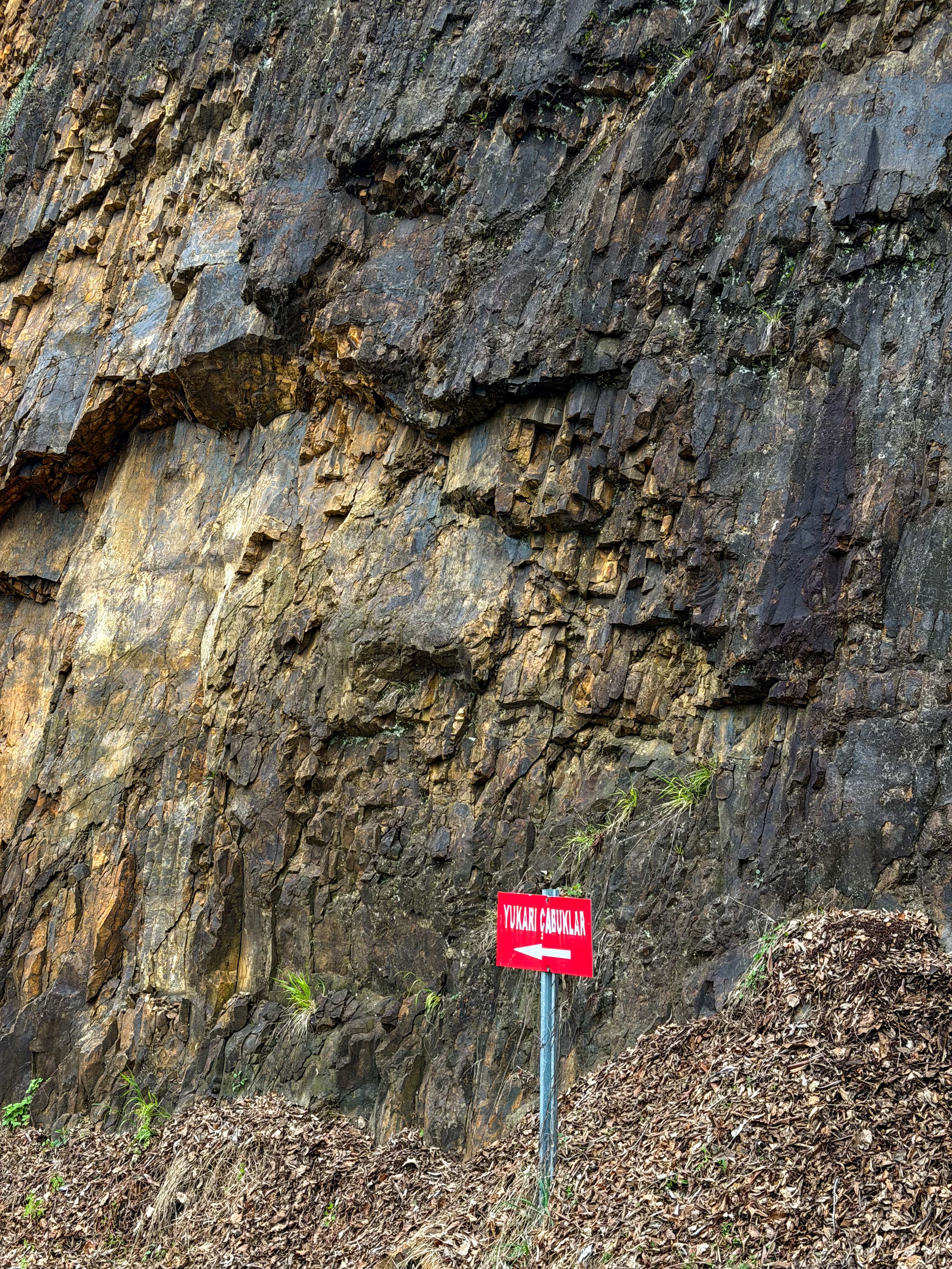 A rugged rocky cliff face with a prominent red caution sign warning of vehicular danger.