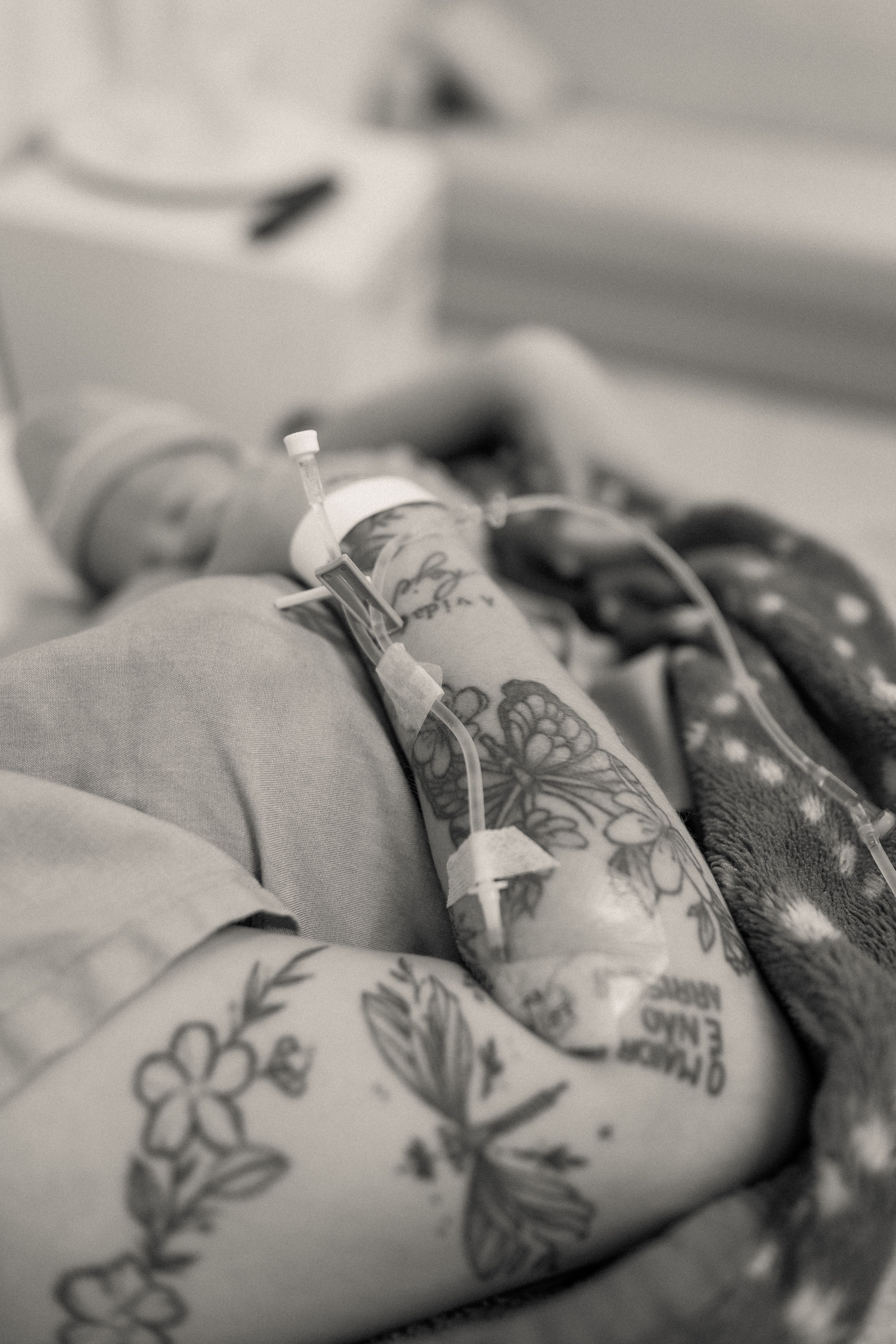 A close-up of a tattooed parent's arm with IV next to a newborn baby in a hospital setting.
