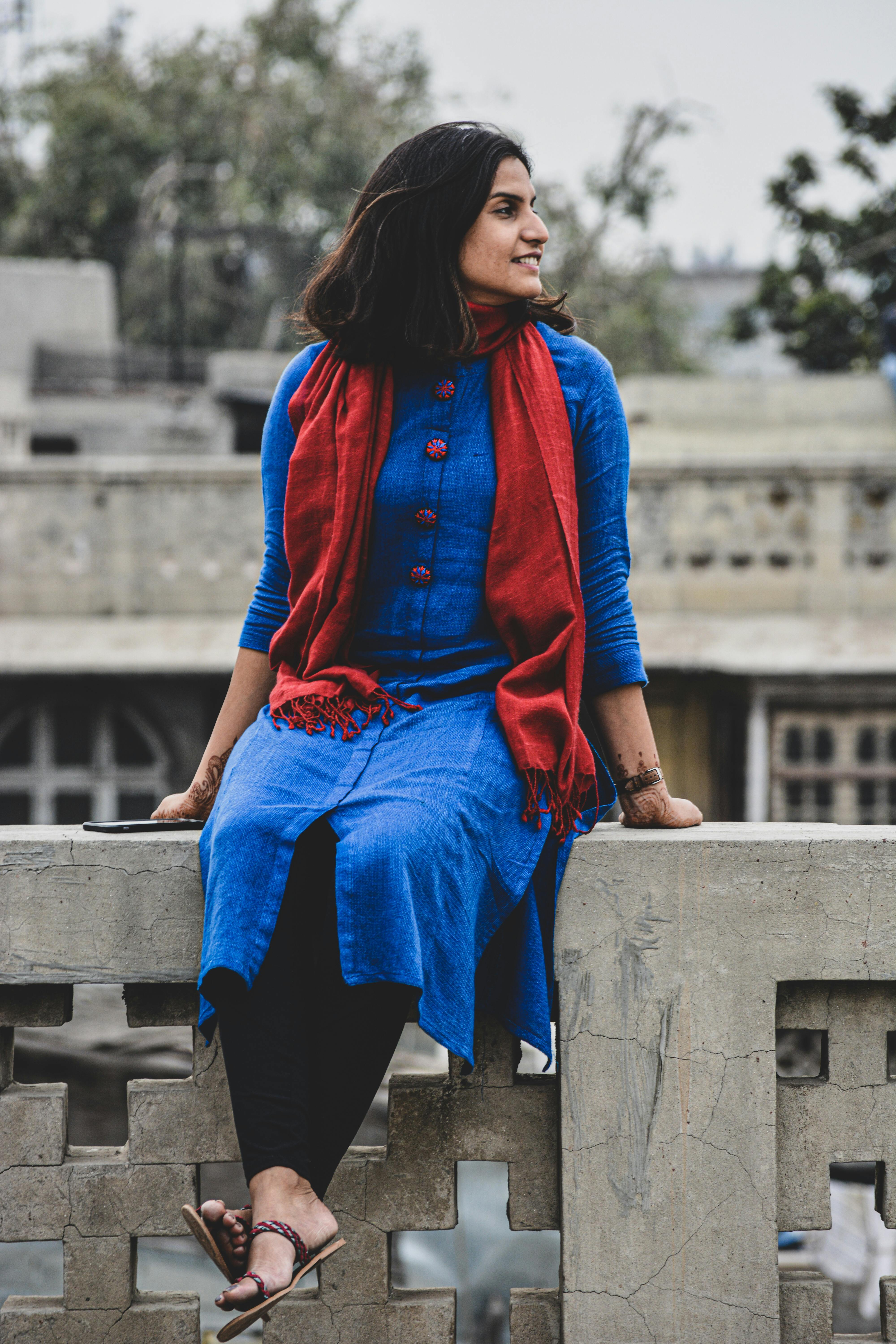 Positive stylish young ethnic female in colorful clothes with mehendi on wrists sitting with crossed legs on concrete fence behind house and trees and looking away