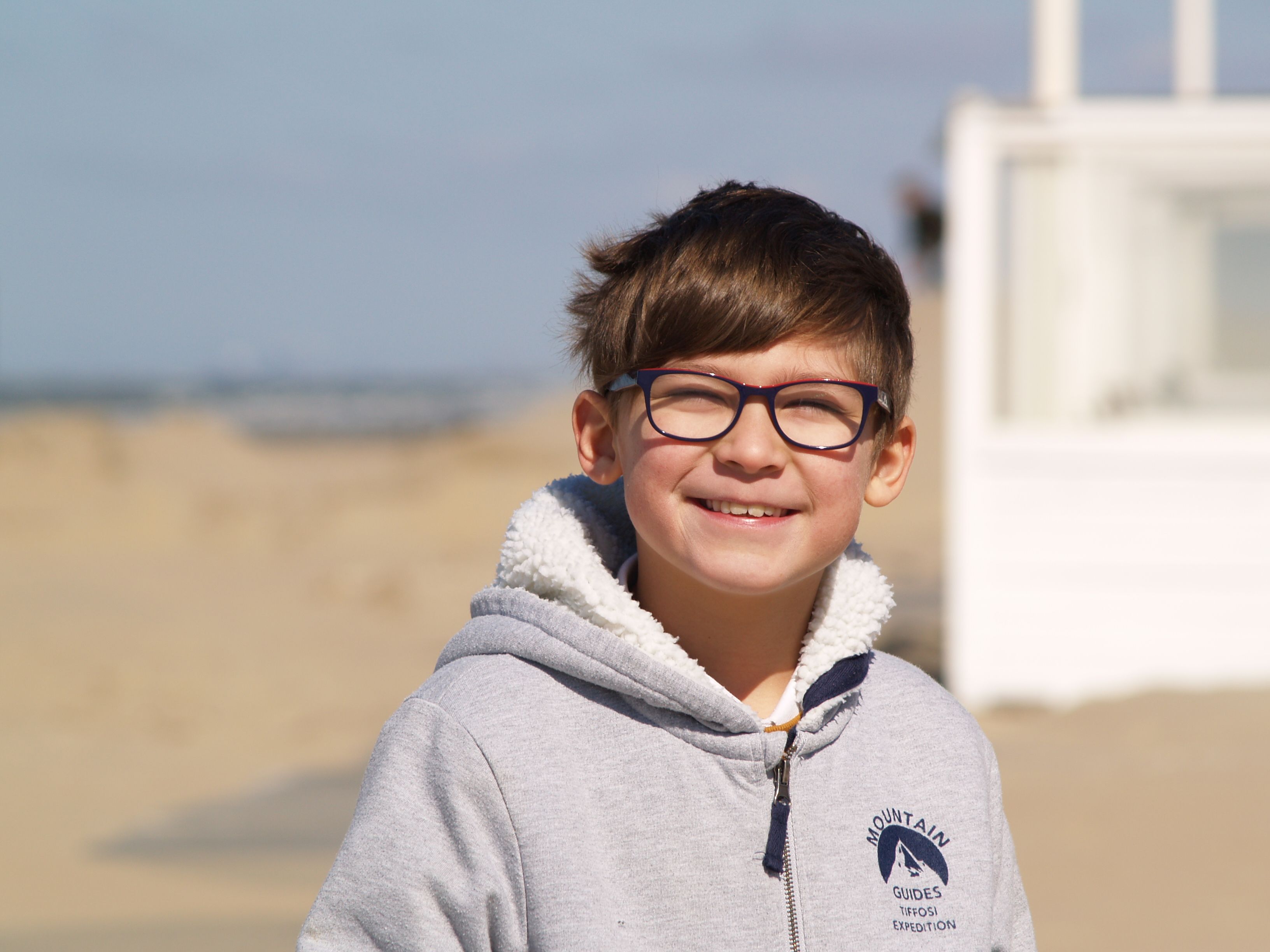 A cheerful child with glasses smiles at the beach, wearing a cozy hoodie on a sunny day.