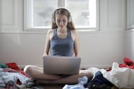 一位年轻女子盘腿坐在床上，戴着耳机使用笔记本电脑