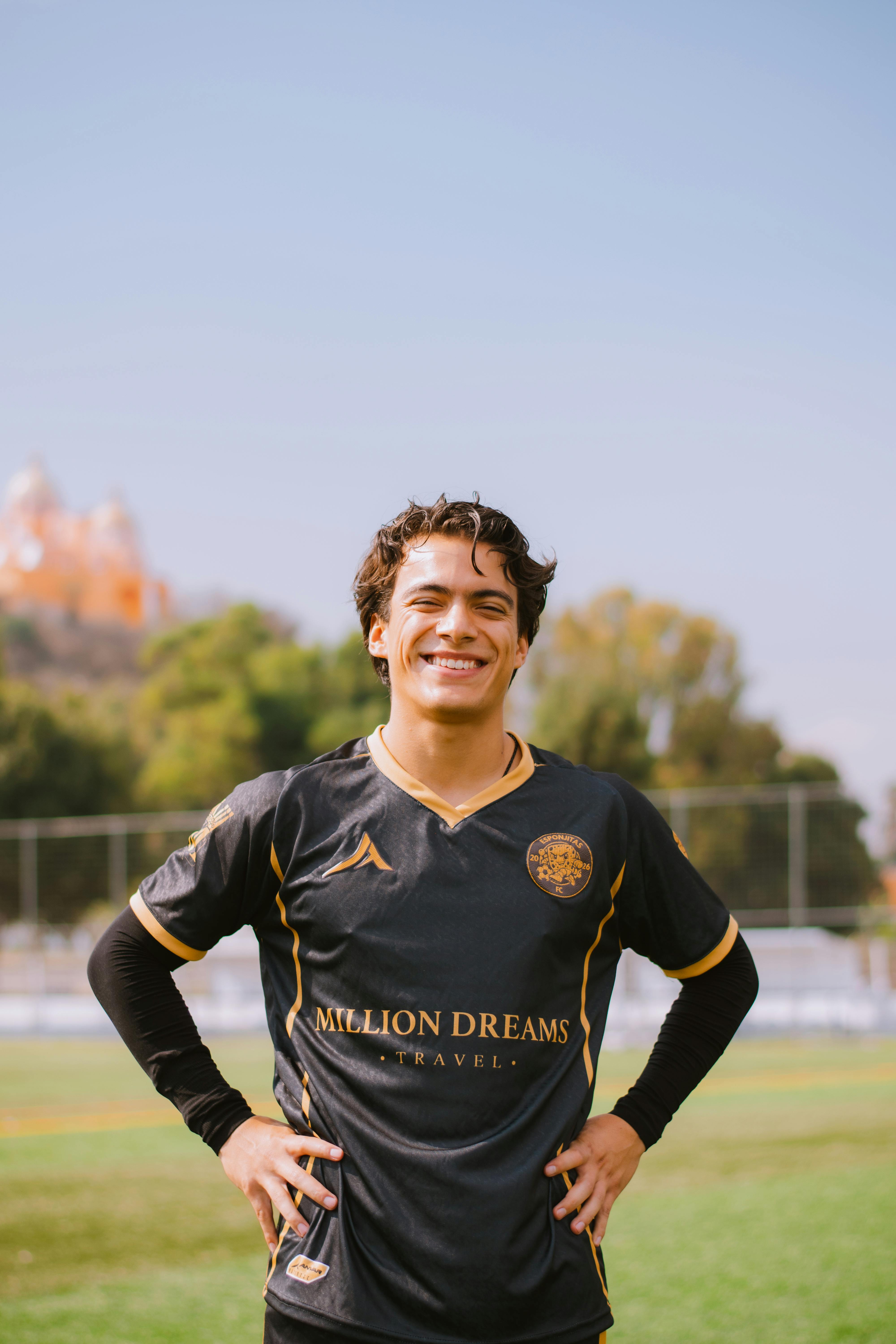 Confident soccer player smiling on a sunny day, wearing a team jersey, with a church visible in the background.