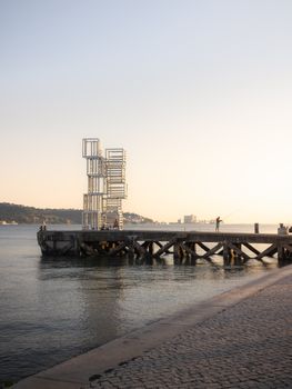 A minimalist steel structure on a pier in Lisbon, captured at sunset over the calm water.