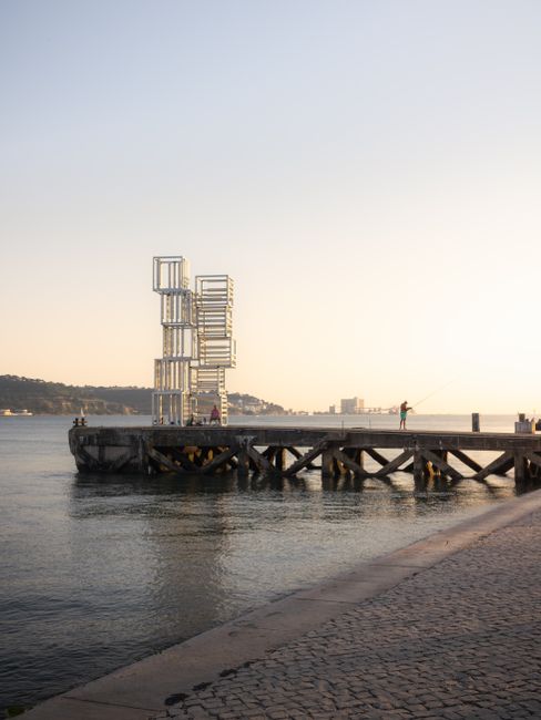 A minimalist steel structure on a pier in Lisbon, captured at sunset over the calm water.