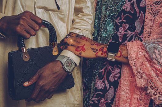 Closeup of hands with watches and henna, dressed in traditional attire, holding a purse.