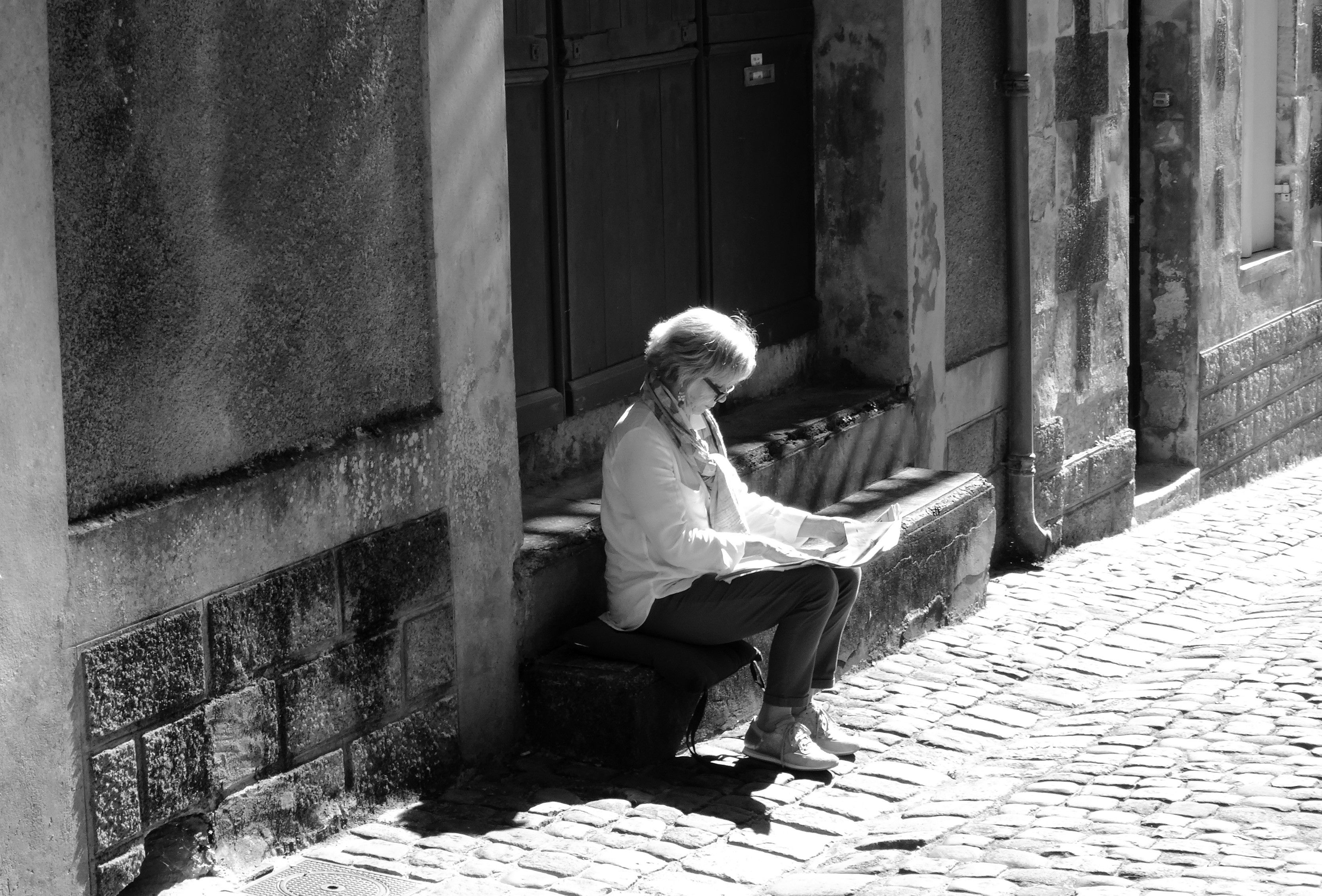 Elderly woman reading in a sunlit cobblestone alley in Saint-Émilion, France.