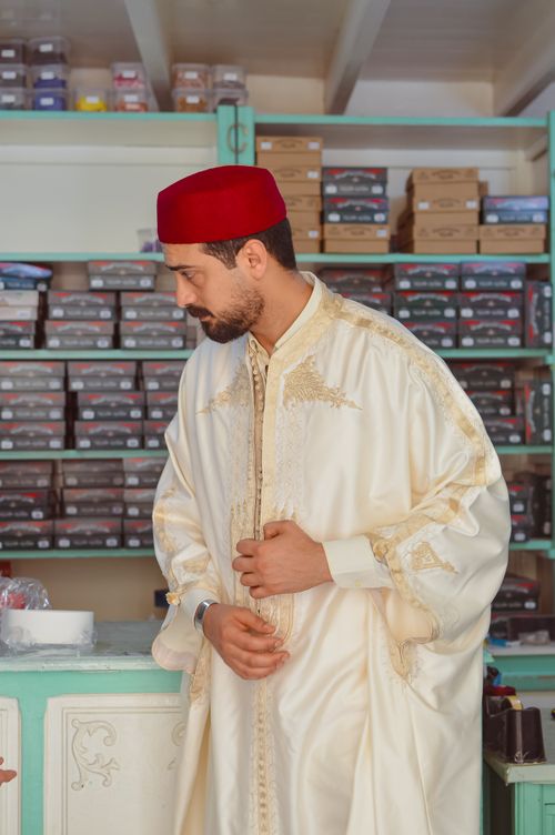 Free A man in traditional Tunisian attire stands inside a medina shop surrounded by boxes. Stock Photo