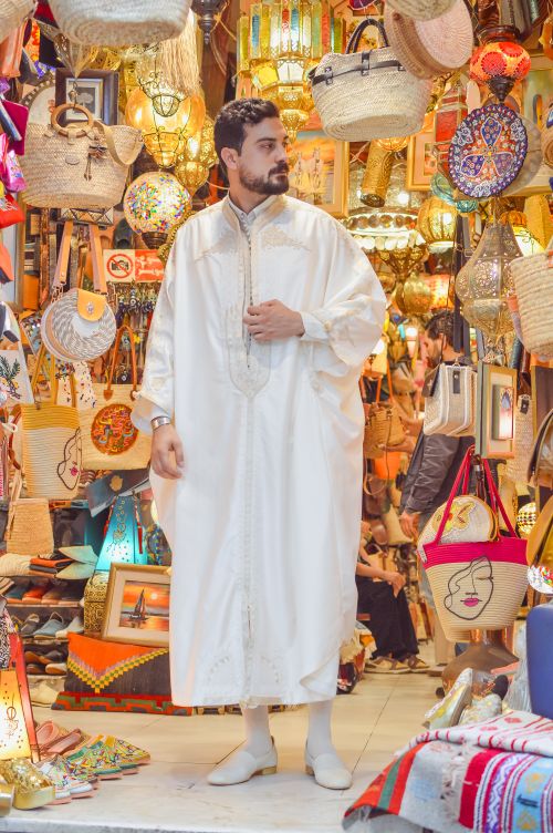 Free Man in traditional attire amidst vibrant market displays of bags and lamps. Stock Photo