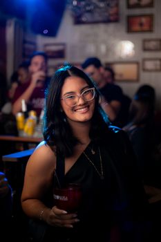 Smiling woman with glasses holding drink at lively indoor bar setting.