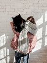 Back view of slim anonymous female in padded jacket and warm scarf cuddling black pet while standing in loft style building in daylight