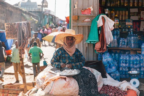 Ethnic male in national clothes and hat looking at camera while sitting on sack with knife and plate full of garlic near product stall and people walking on old street