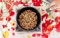 Top view of crop unrecognizable cook frying halved brown and golden crunchy peanuts on old portable gas stove while holding handle of frying pan above colorful oilcloth on table