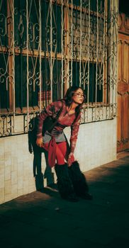 Fashionable woman in fur boots leaning against an ornate iron fence in sunlight.