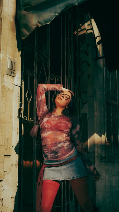 Free Young woman in stylish attire poses in sunlight with rustic gate backdrop, embodying urban fashion with artistic flair. Stock Photo