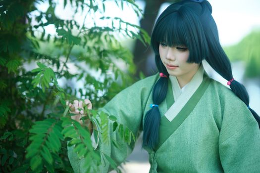 Young woman in green traditional attire, admiring foliage outdoors on a sunny day.