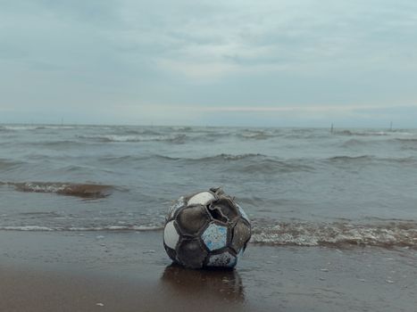 A worn-out soccer ball lies on the sandy beach with waves approaching.