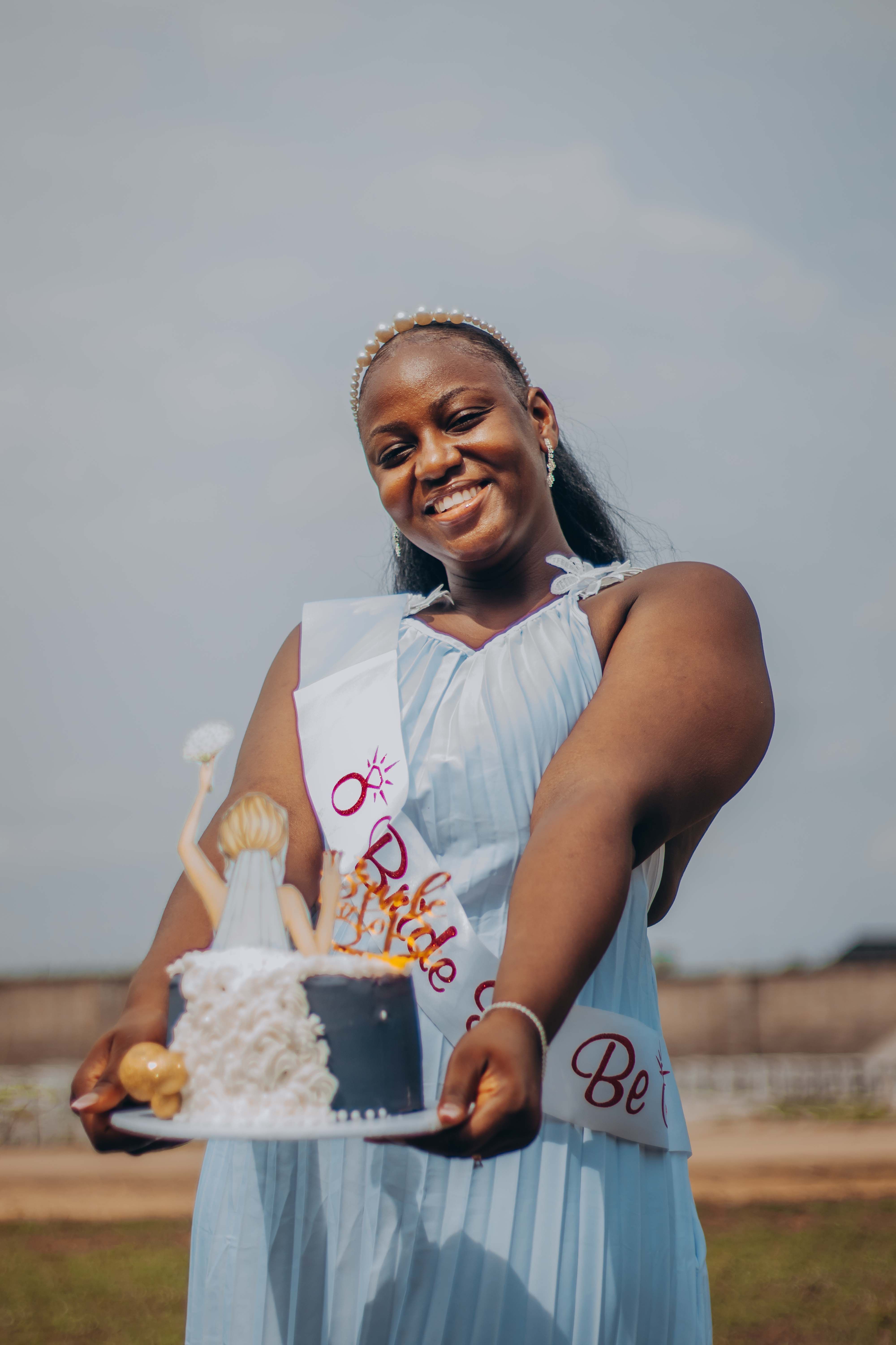 Joyful bride-to-be holds decorative cake wearing a sash, celebrating outdoors under a clear sky.