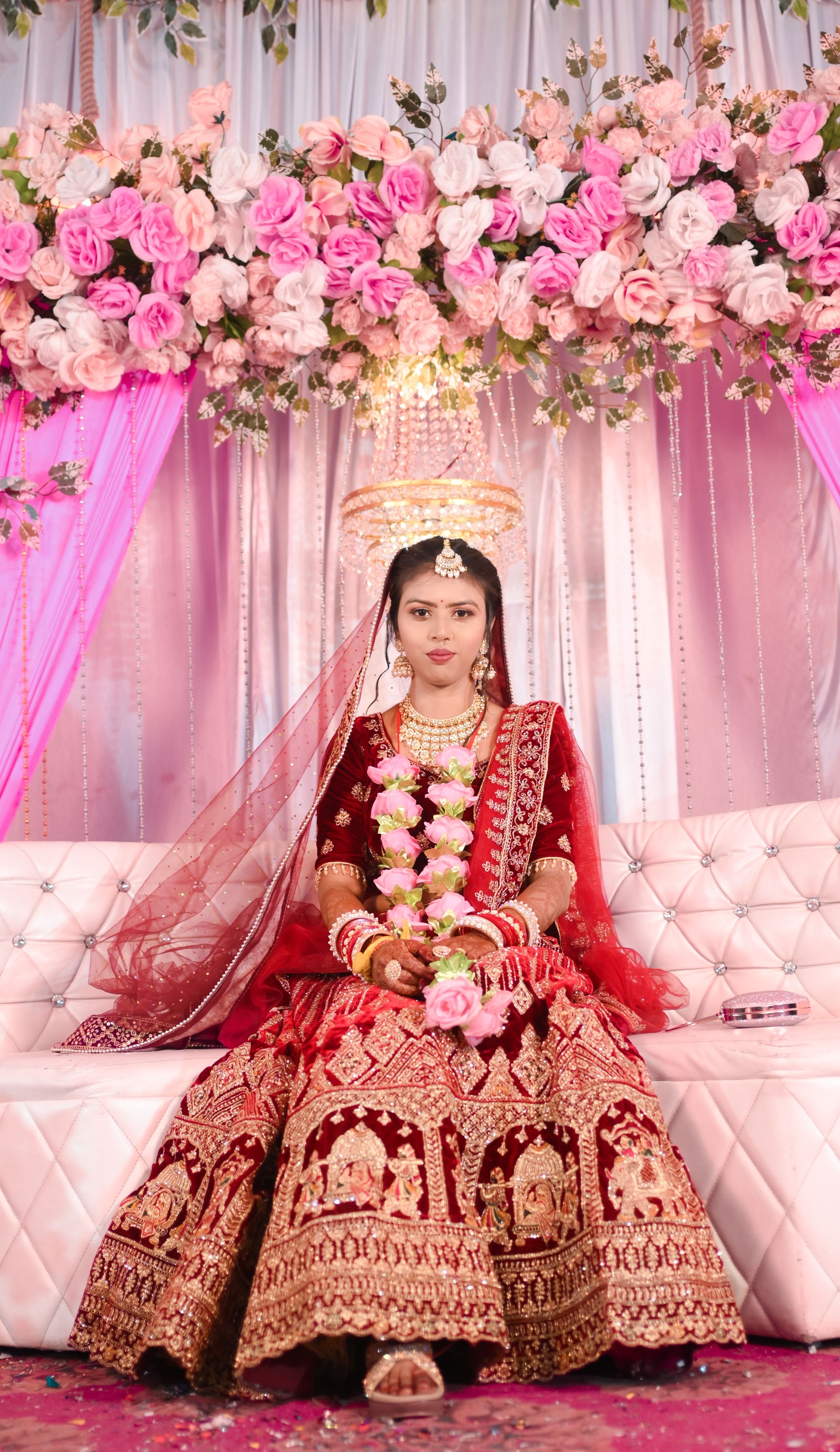 Elegant bride in red traditional attire seated on a decorated couch during a wedding.
