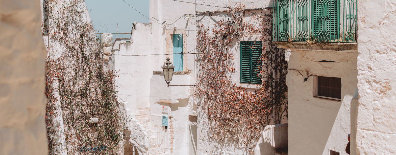 Explore the picturesque alleys of Ostuni, Apulia, with sunlit whitewashed buildings and green shutters.