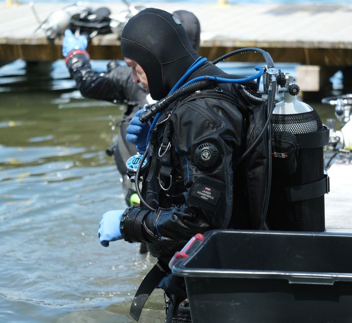 Divers in wetsuits and scuba gear on a dock, adjusting masks and tanks before entering the water.