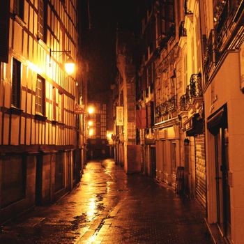A quiet, wet street in Bayonne, France, glows under city lights at night. Reflective pavement creates an atmospheric scene.