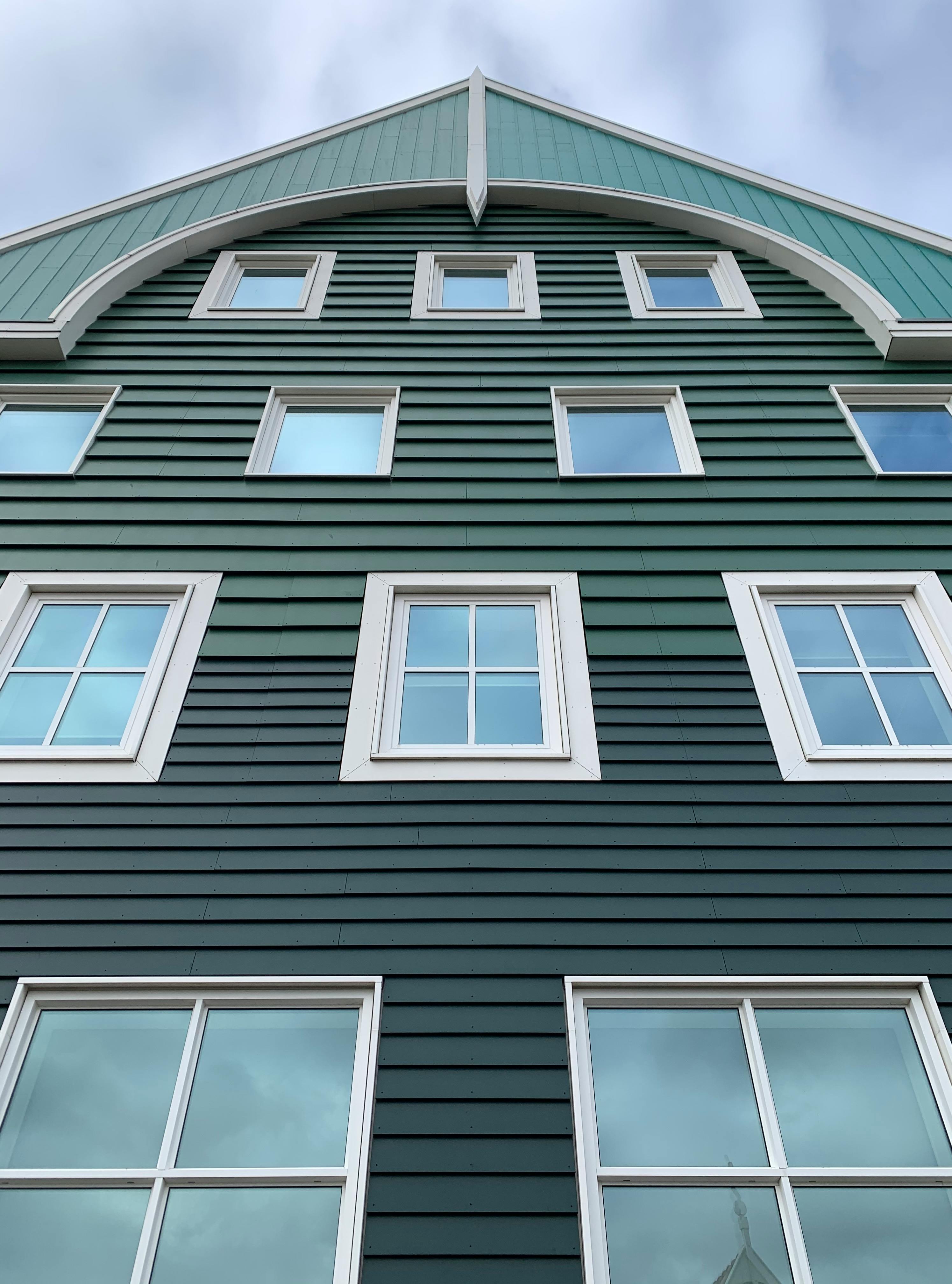 Green wooden building with white-framed windows under a teal roof against a cloudy sky.
