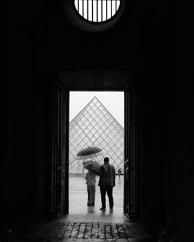Black and white image of a couple with an umbrella standing near the iconic Louvre Pyramid in Paris.