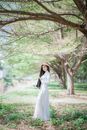 一位身着白色连衣裙、头戴遮阳帽的女子，优雅地站在茂密的树枝下