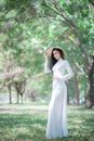 一位身穿白色连衣裙、头戴太阳帽的美丽亚洲女子，优雅地站在郁郁葱葱的绿色公园里