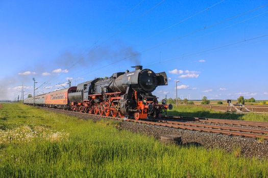 A vintage steam locomotive travels through picturesque countryside under a clear blue sky.
