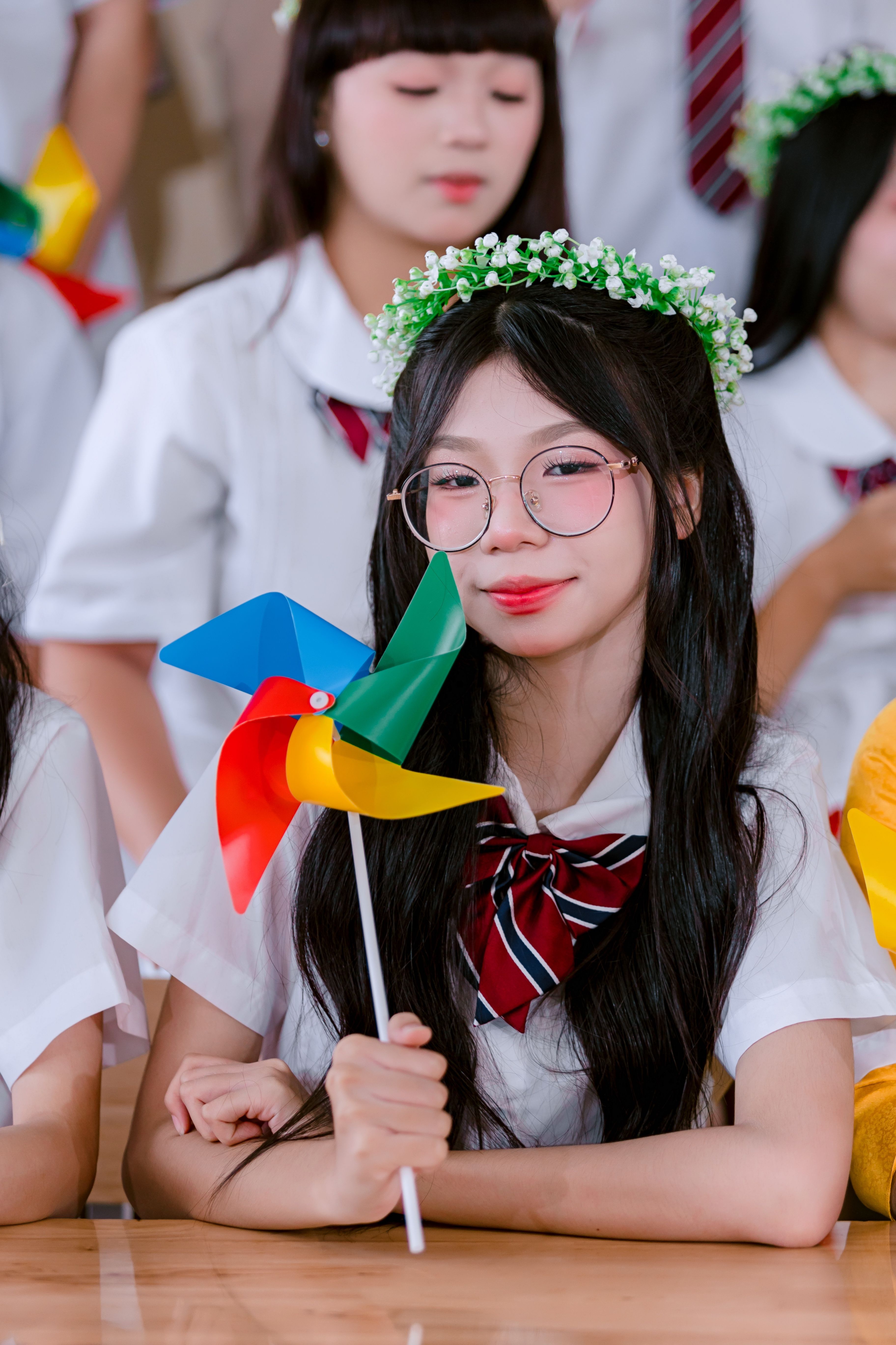 Cute teen girl with glasses holding a colorful pinwheel indoors, wearing a uniform and floral headband.